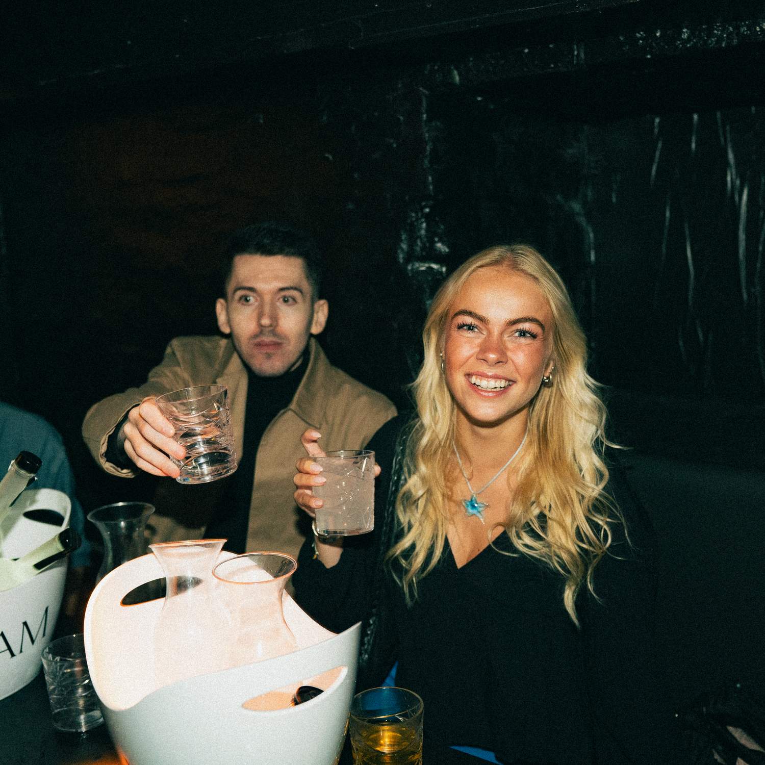 Two people at a dimly lit bar enjoying drinks together as they smile at the camera.