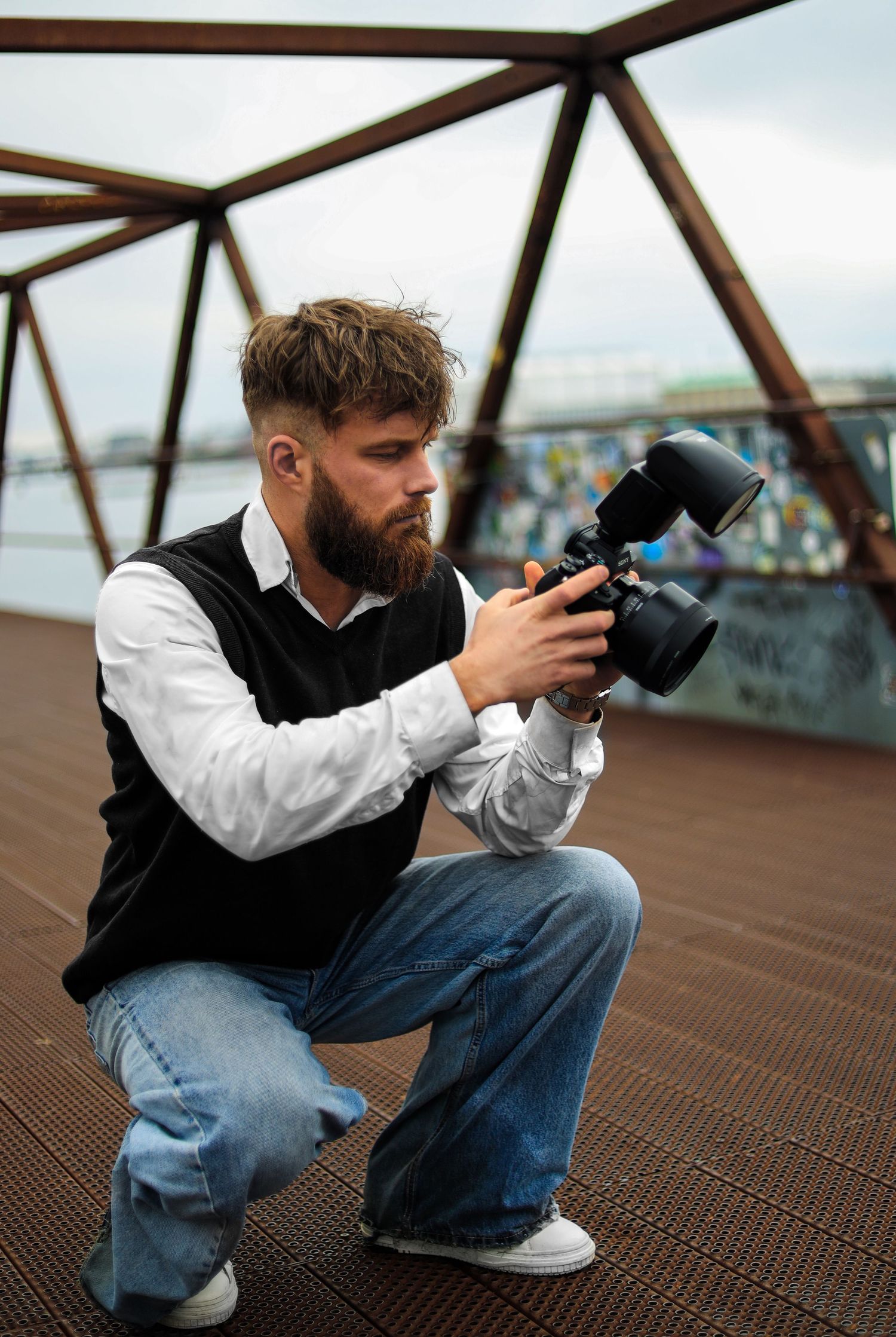 Professional photographer crouching on rusty bridge deck adjusts camera settings while wearing vest and white shirt.