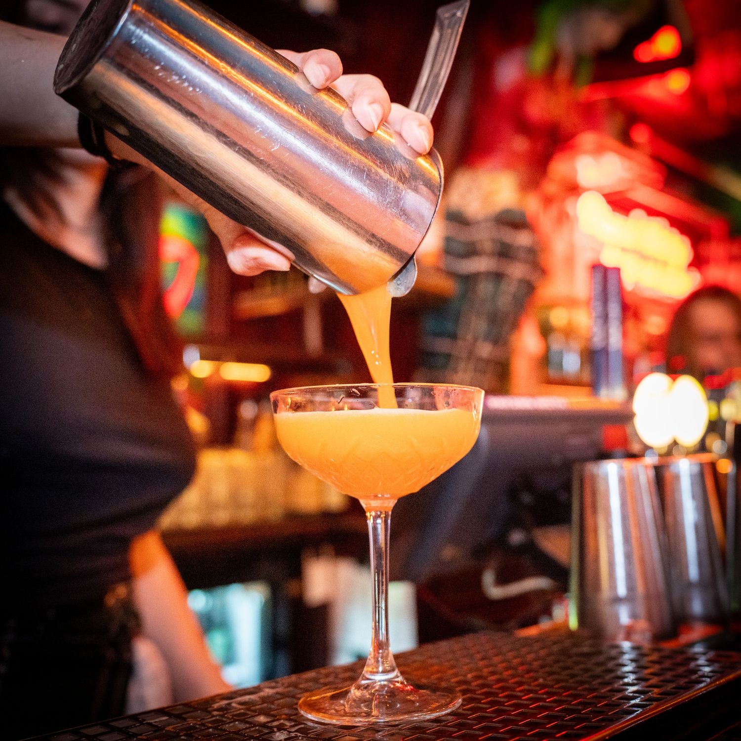 Bartender hælder en gul cocktail fra en shaker i et coupeglas på en oplyst bar.