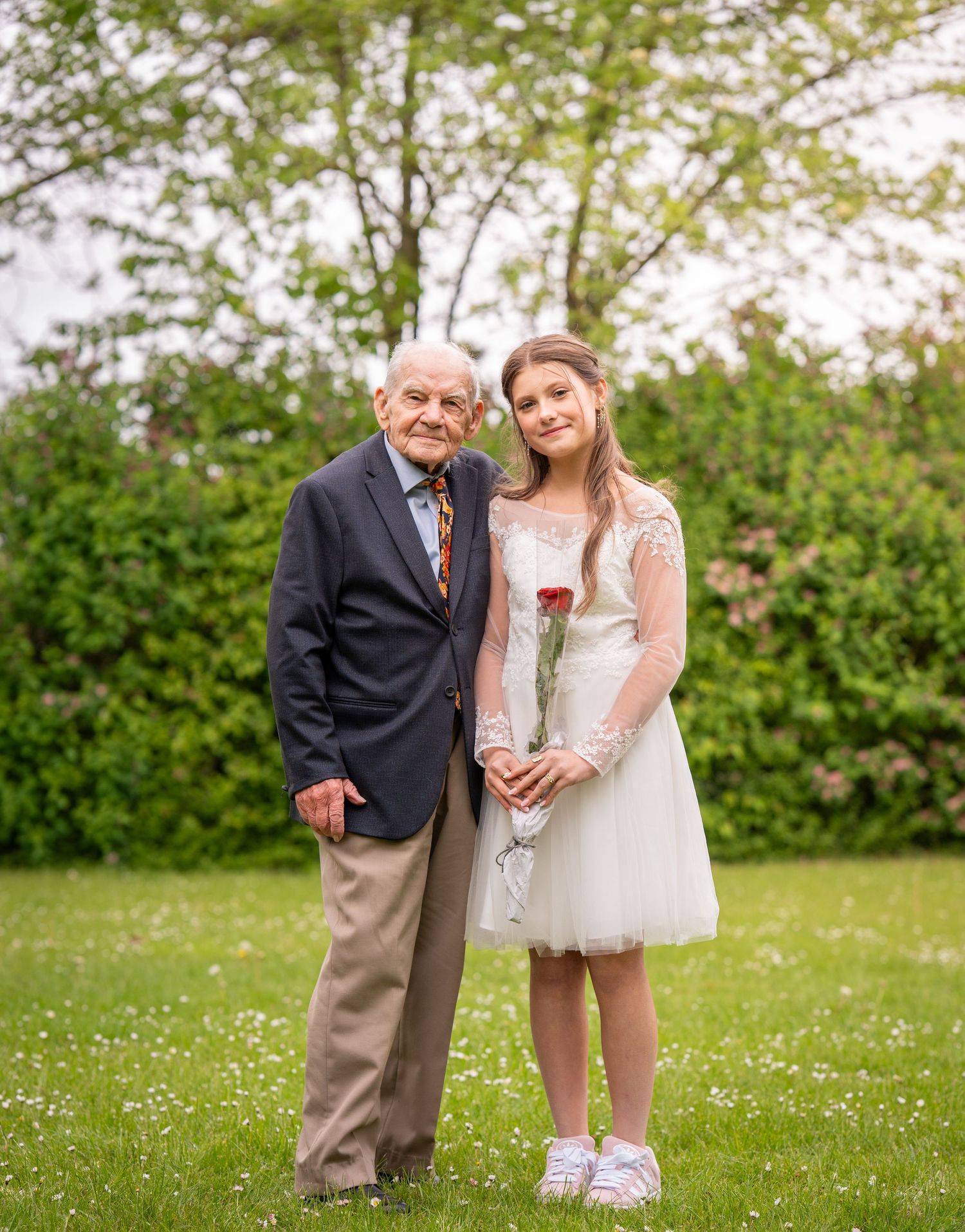 A pair stand together on a grassy lawn with spring trees in the background, one holding a rose.