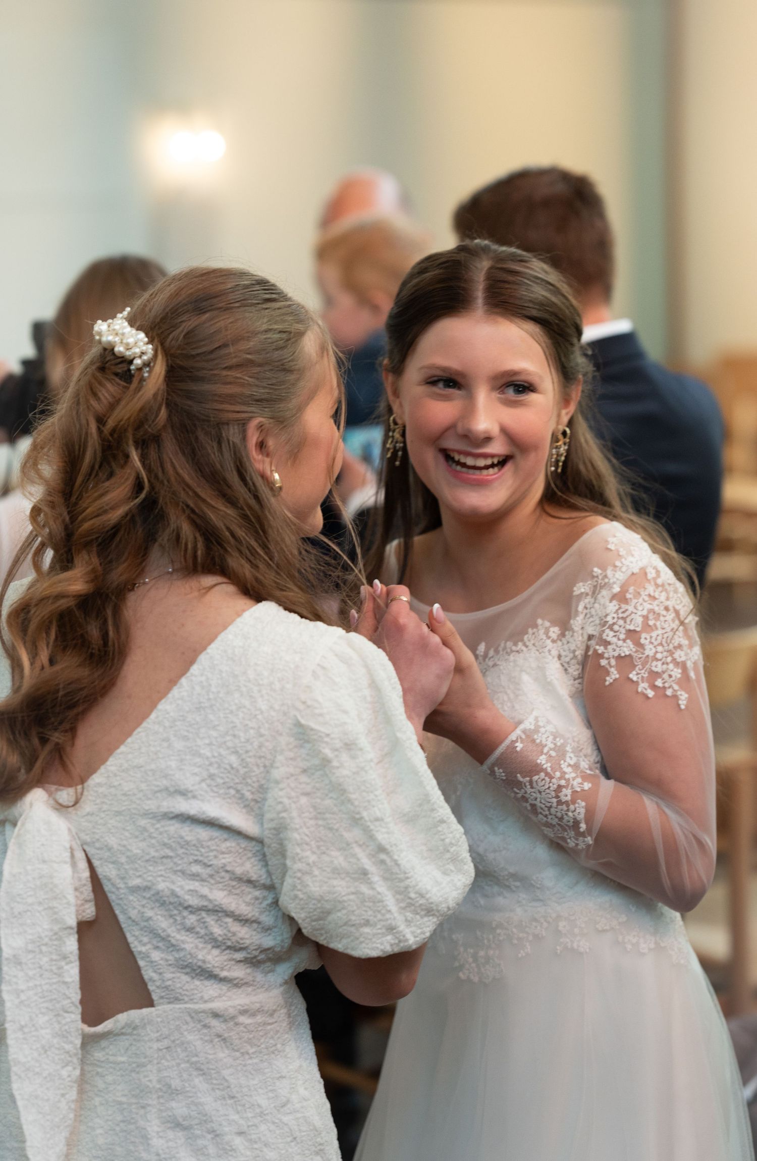 Two people in elegant white lace wedding dresses share a joyful moment while conversing at a celebration.