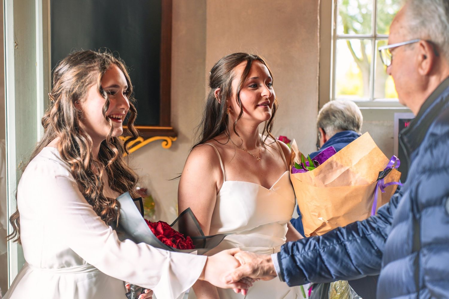 A joyful moment shows two people in wedding attire greeting someone in blue while receiving a small gift.