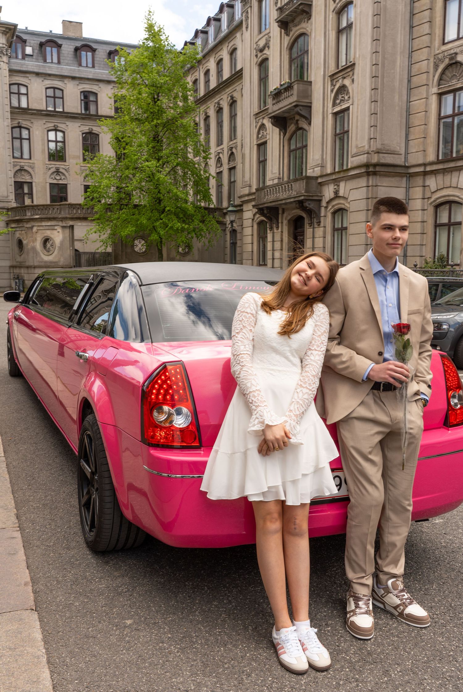 A couple poses next to a bright pink stretch limousine on a European street with historic buildings in the background.