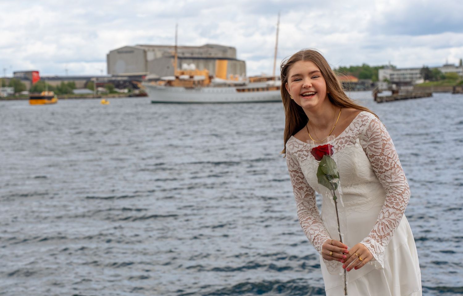 Someone wearing a cream-colored lace dress holds a red rose while standing on a waterfront with industrial buildings behind them.