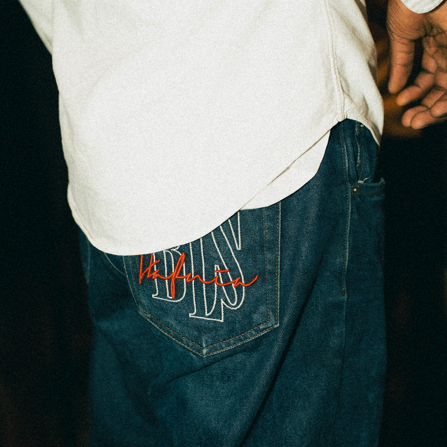 Close-up view of hip pocket detail on dark denim jeans with red embroidered branding against black background.