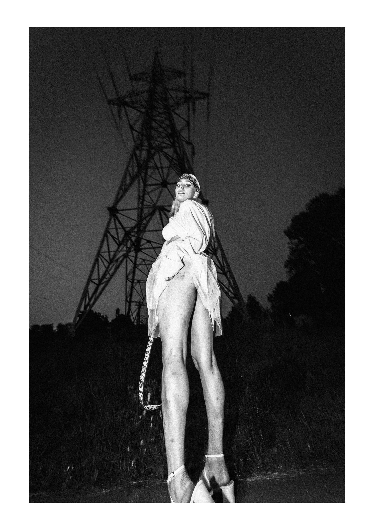 Black and white artistic photograph of a figure in flowing white fabric posing dramatically near an electrical tower at night.