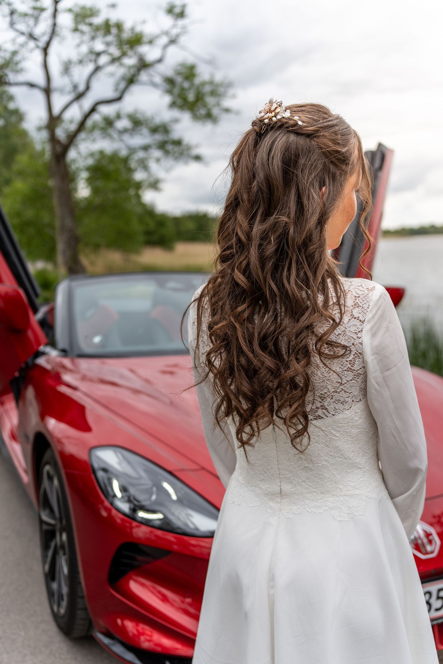 Long curled brown hair adorned with a delicate tiara shown from behind next to a red luxury sports car by a lake.