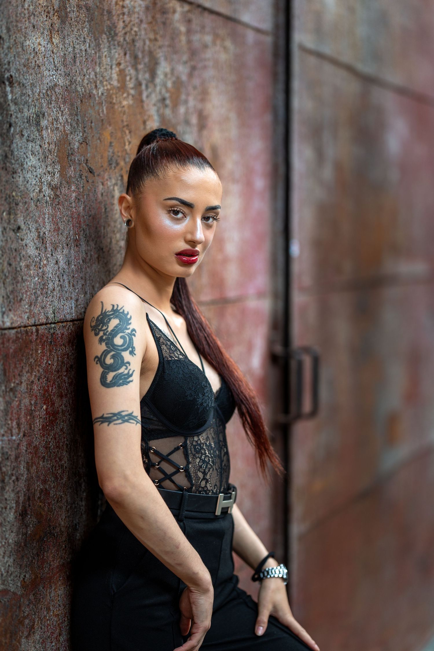 Model in black dress poses against rustic brick wall showing tattoo artwork in moody lighting.