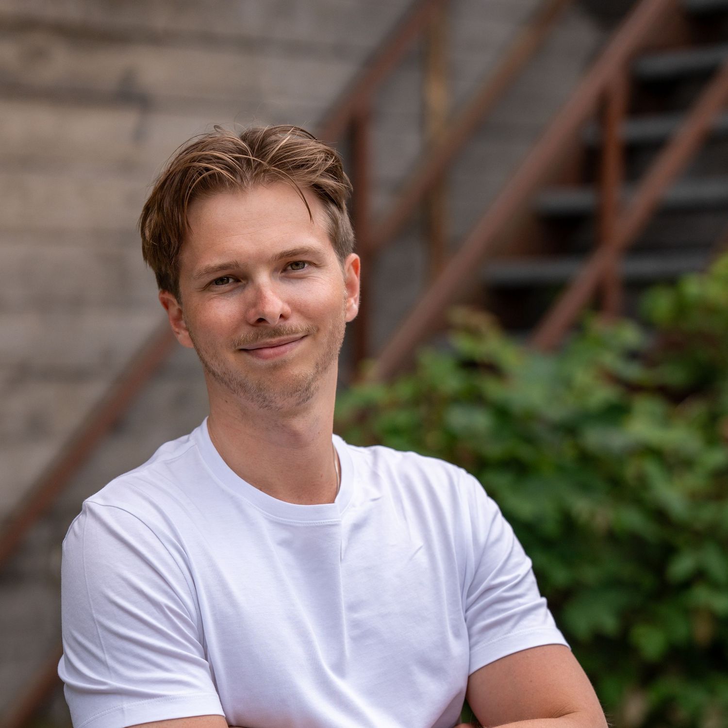 A person in a white t-shirt posing against a rustic wooden background with greenery visible.