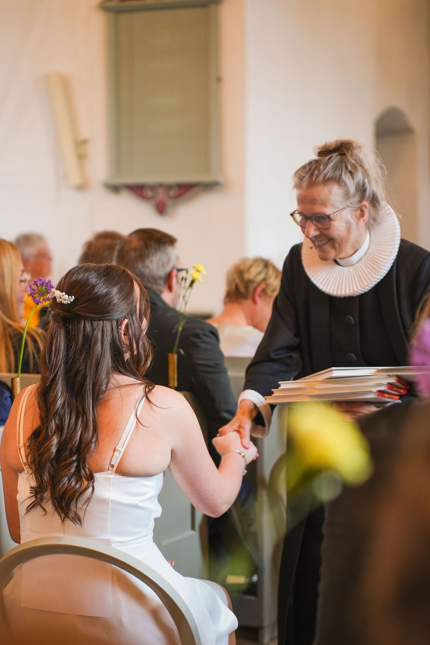 Member of clergy in traditional black robe and white collar speaks with a bride at a wedding ceremony.