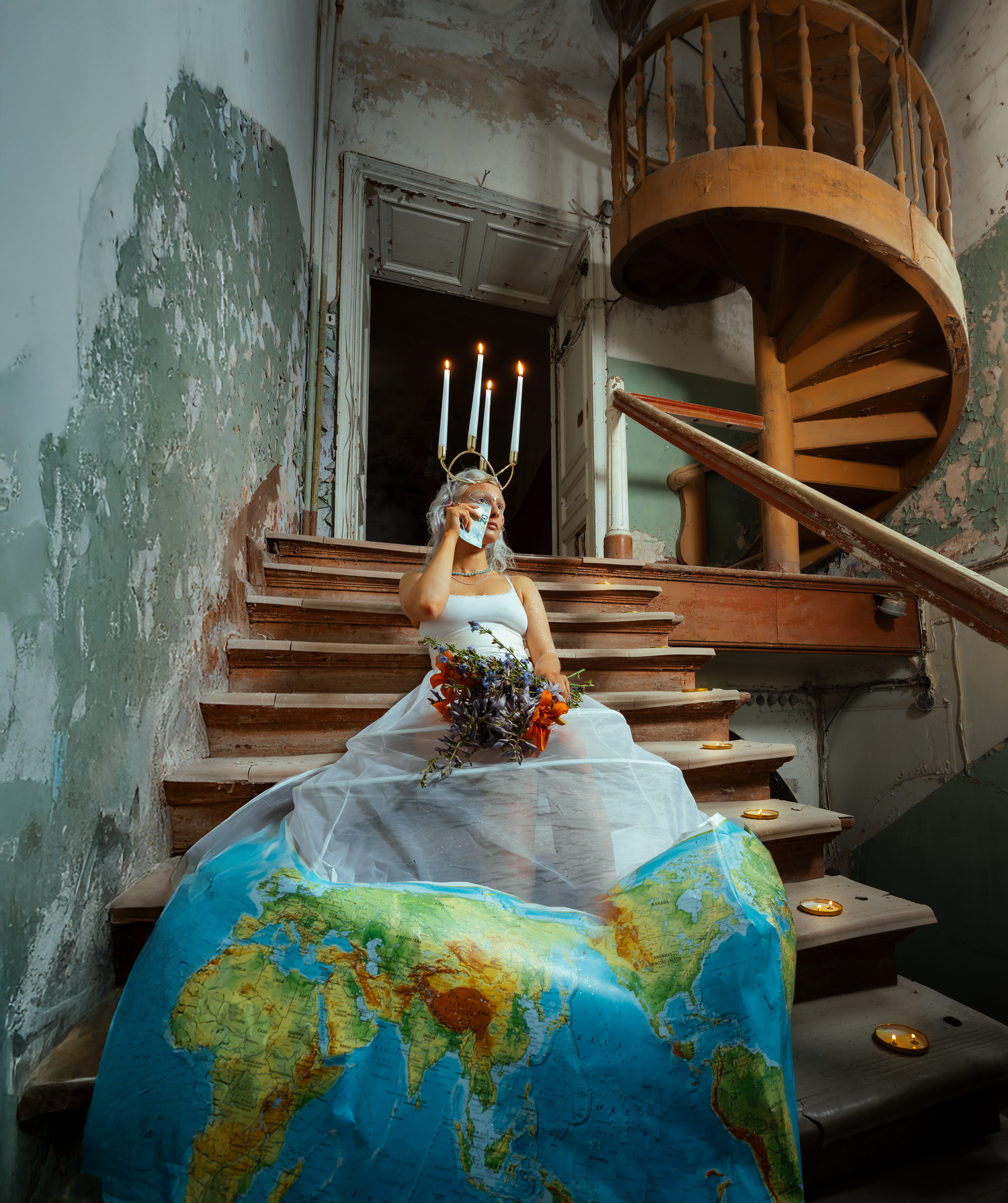 A bride in white dress sits on a world map dress train on vintage wooden spiral staircase in abandoned building.