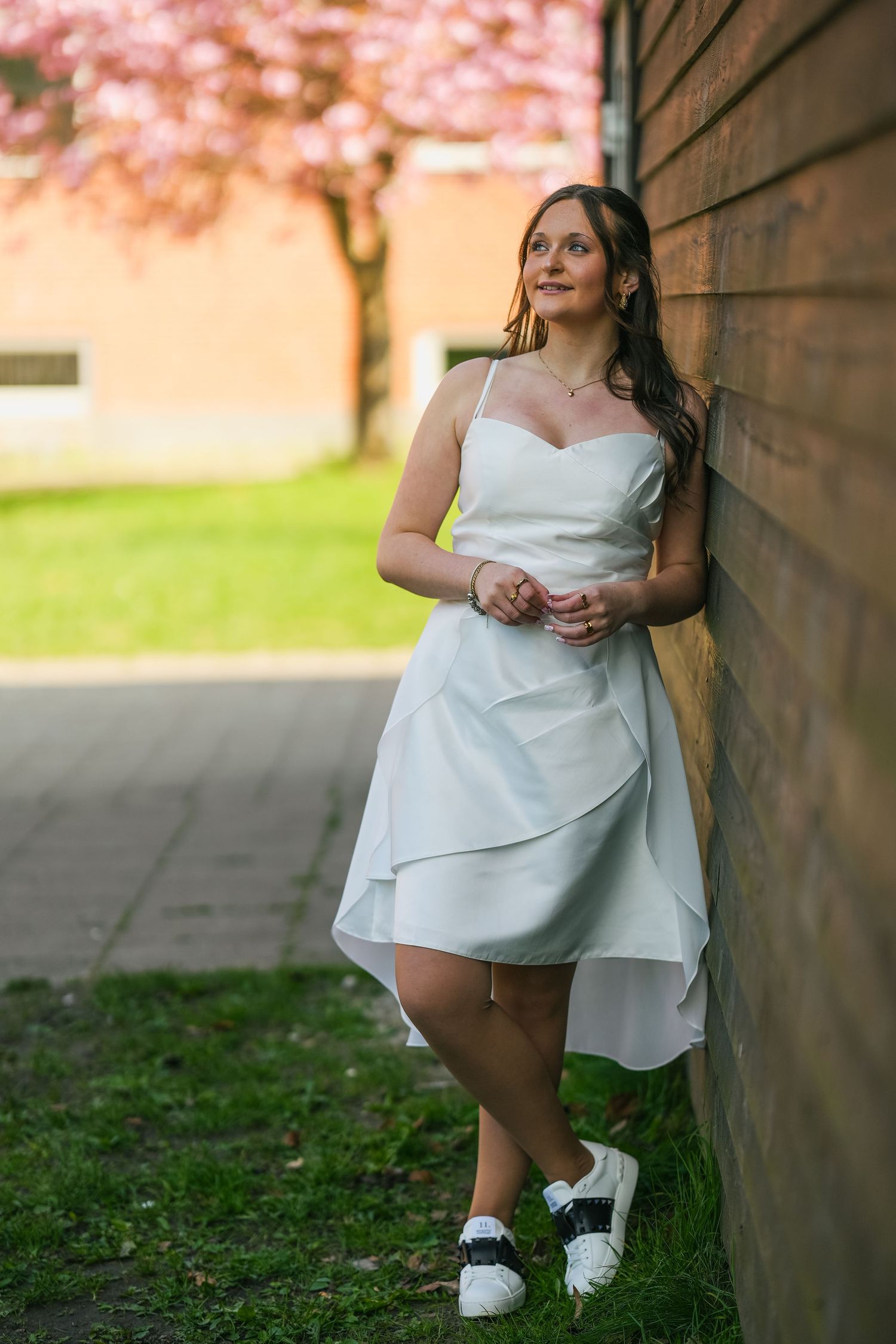A person in a white dress and sneakers leans against a brick wall with cherry blossoms blooming in the background.