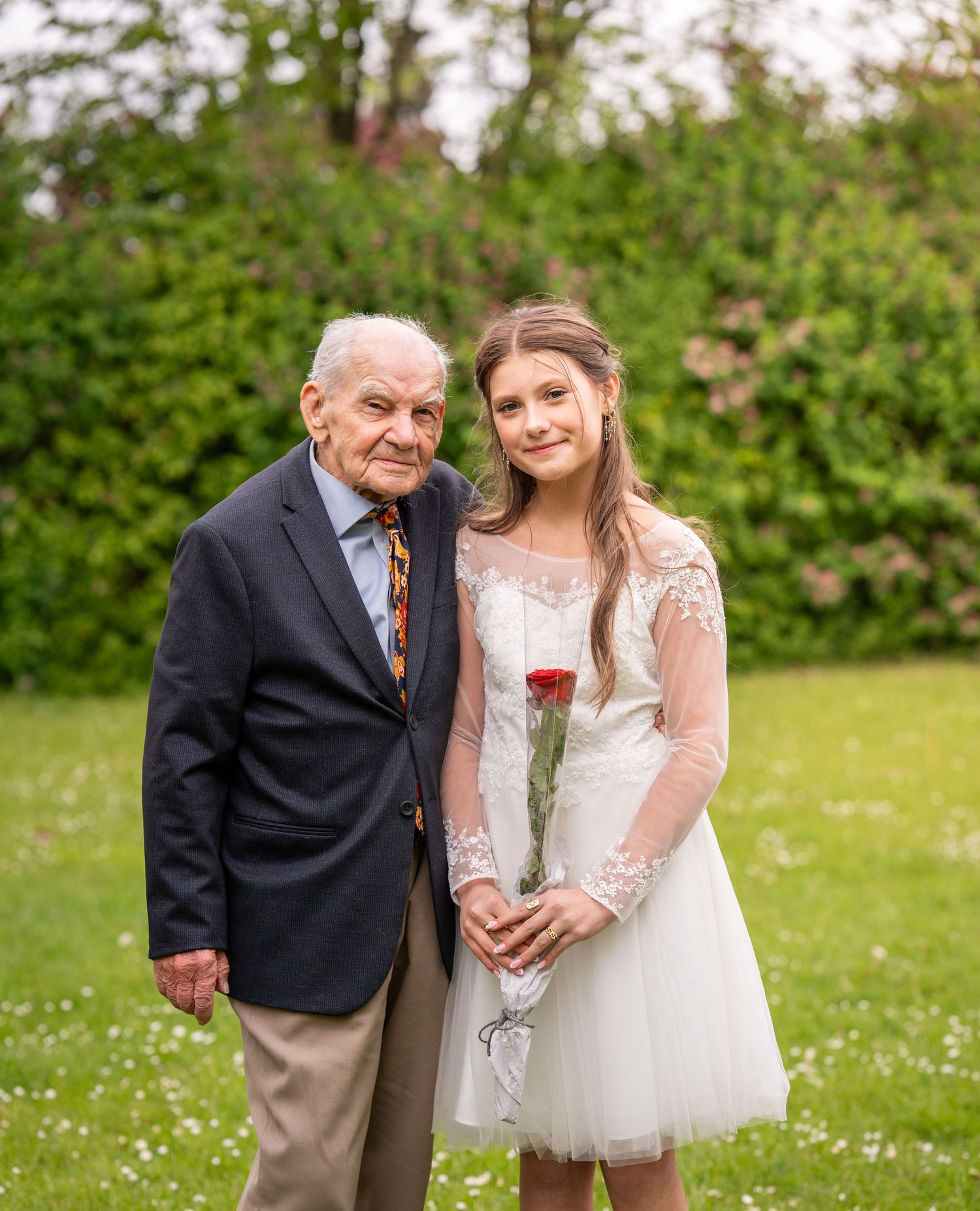 A senior man in a suit and younger person in a lace white dress pose together outdoors holding a red rose.