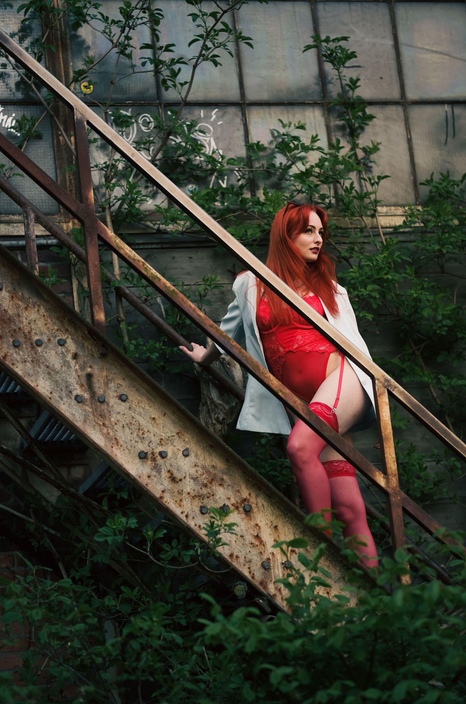 Figure in red dress and jacket poses on rusty metal stairs surrounded by lush green foliage in moody outdoor setting.