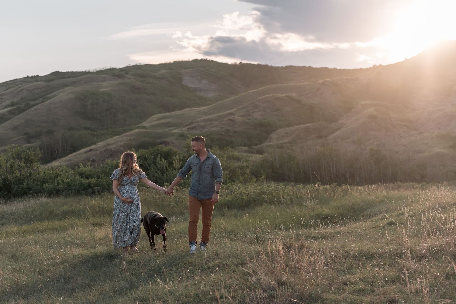 A couple holding hands with their dog in a scenic mountain meadow at sunset with golden light illuminating the landscape.