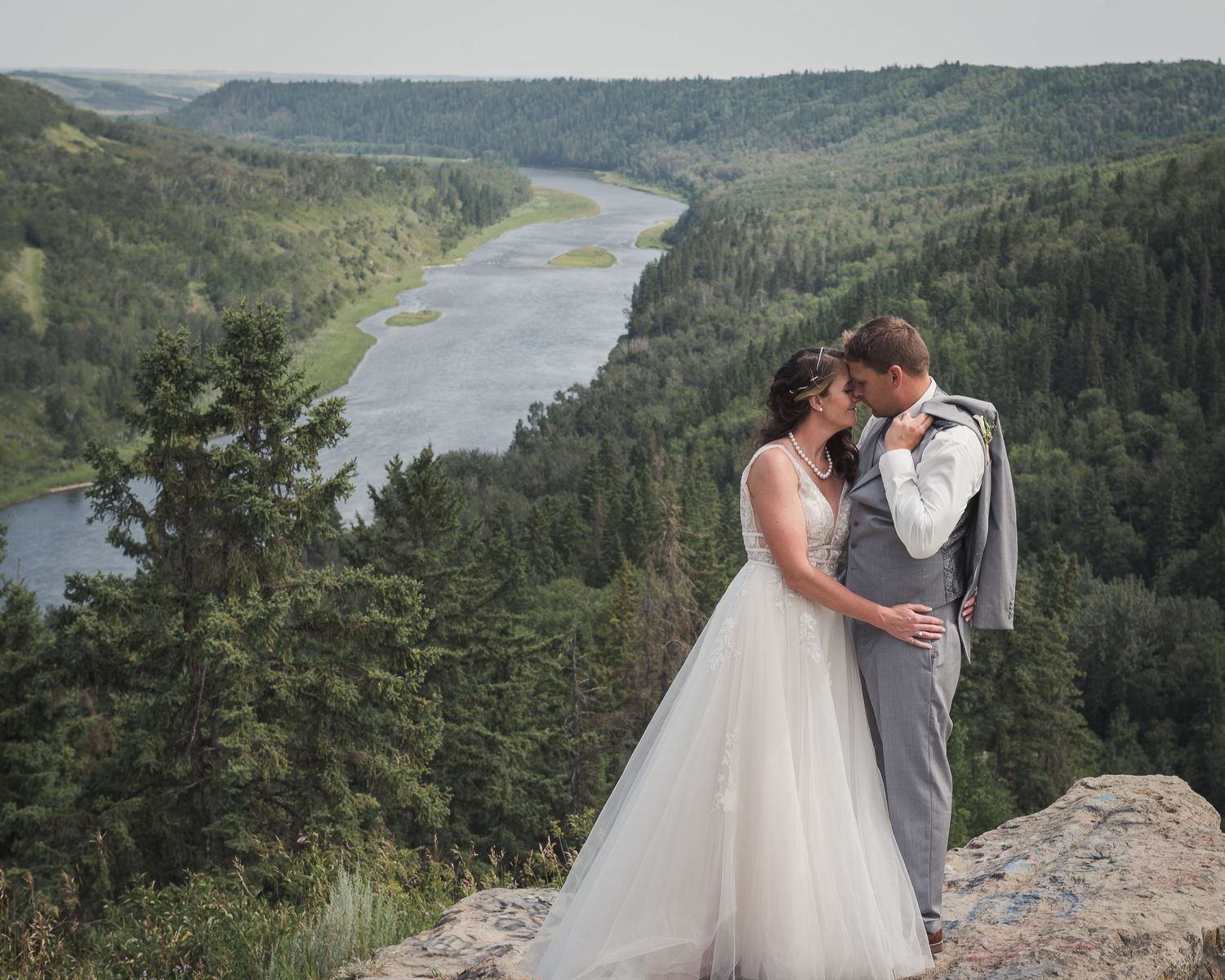 Wedding couple embracing on clifftop overlook with scenic river valley and forested hills in background.