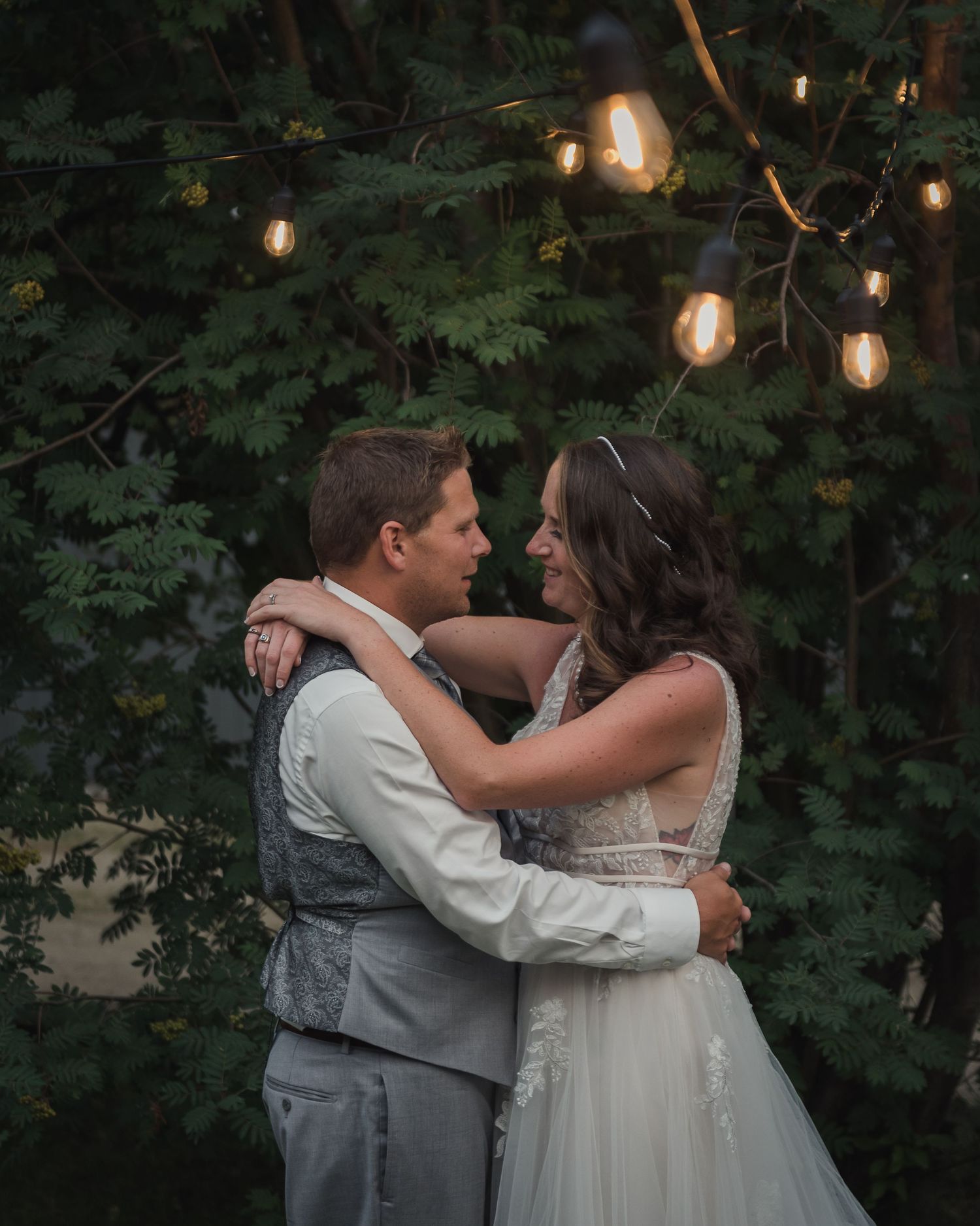 Intimate wedding scene with hanging Edison bulbs illuminating couple dancing in garden at dusk.