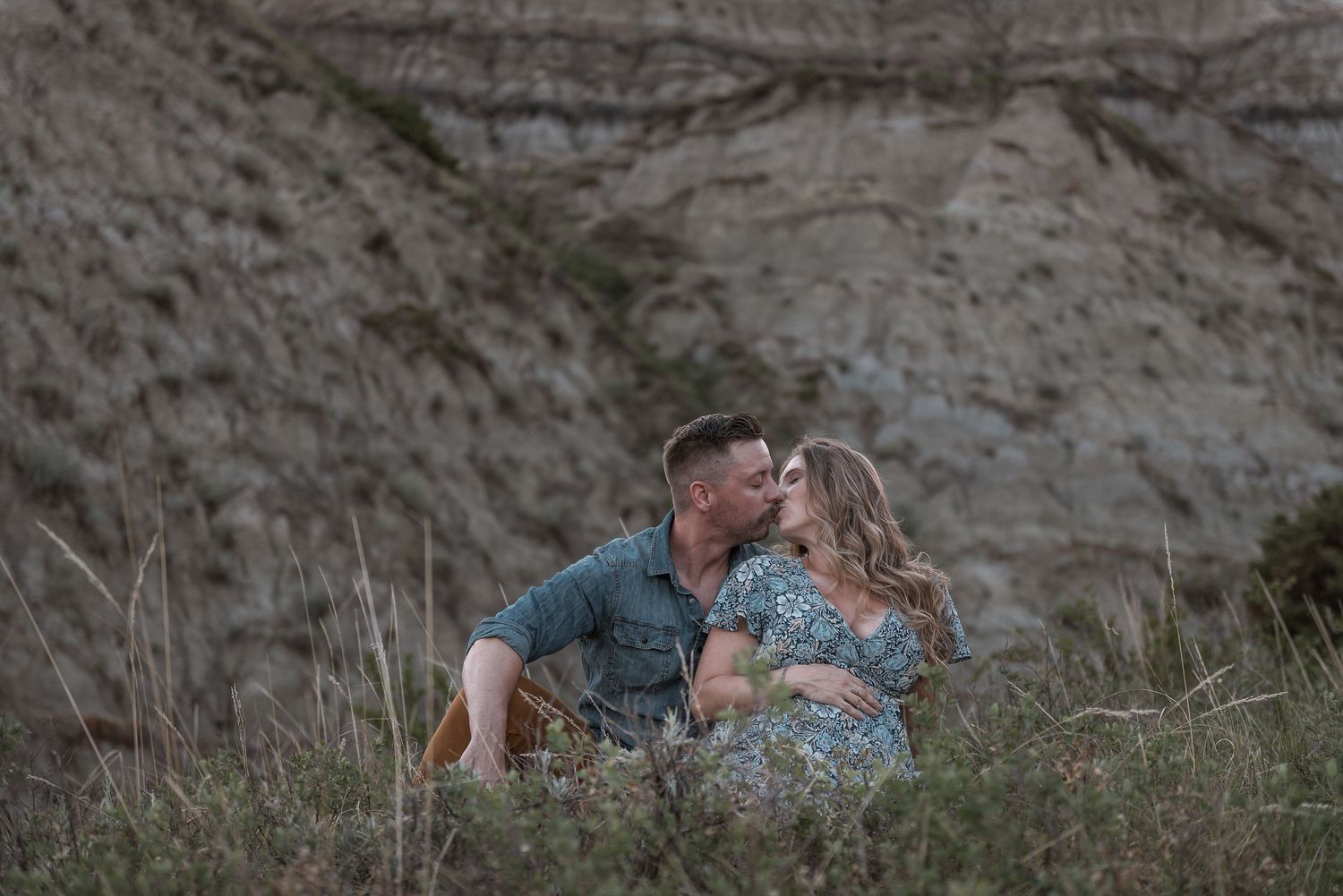 A couple shares a tender moment while sitting in tall grass against a rocky hillside backdrop.