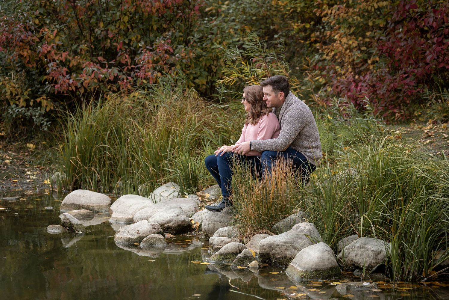 Couple sitting together on rocks by serene pond surrounded by autumn foliage.