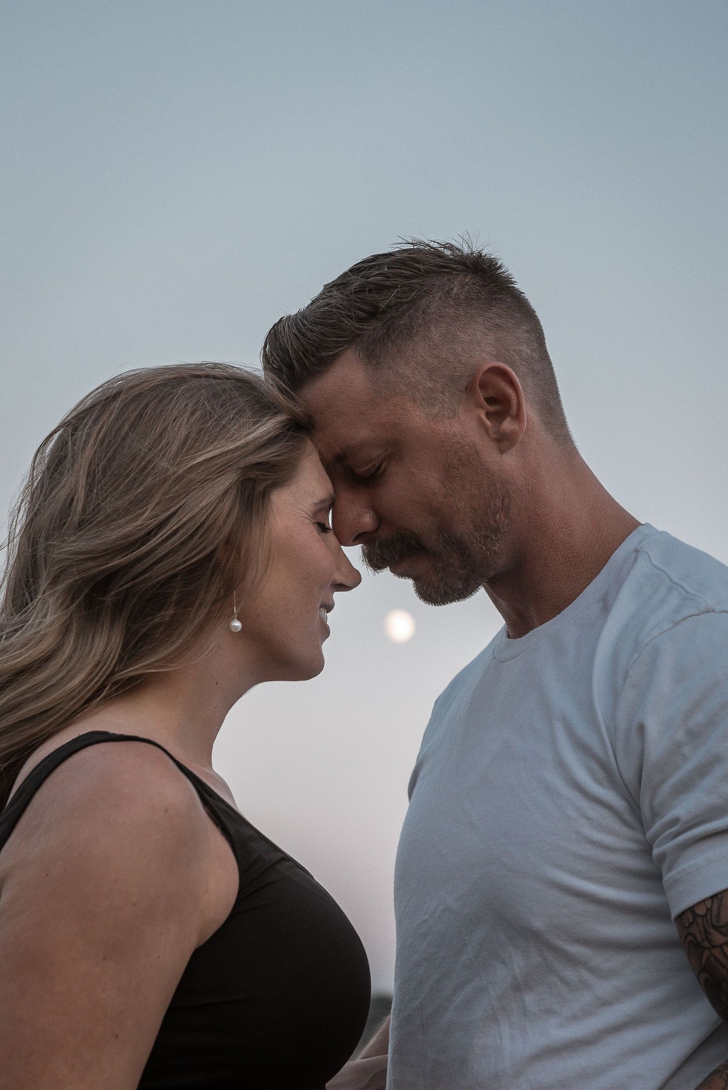 An intimate couple shares a tender moment at dusk, their foreheads touching against a muted sky.