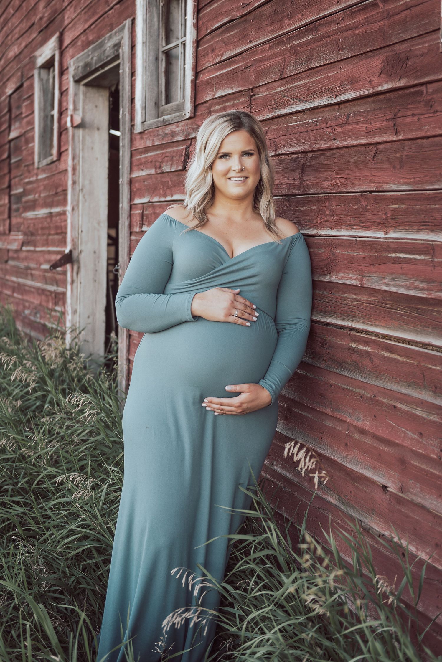 A maternity photo of a woman in a teal dress against a rustic red barn.