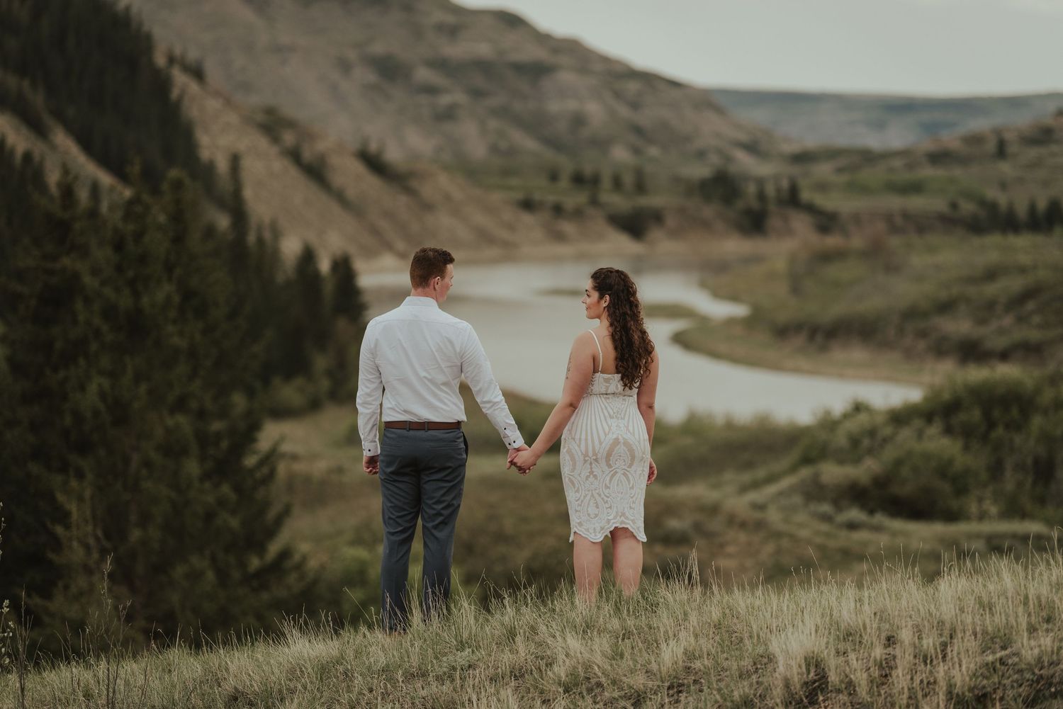 Couple standing hand in hand on scenic overlook with winding river and mountains in background.
