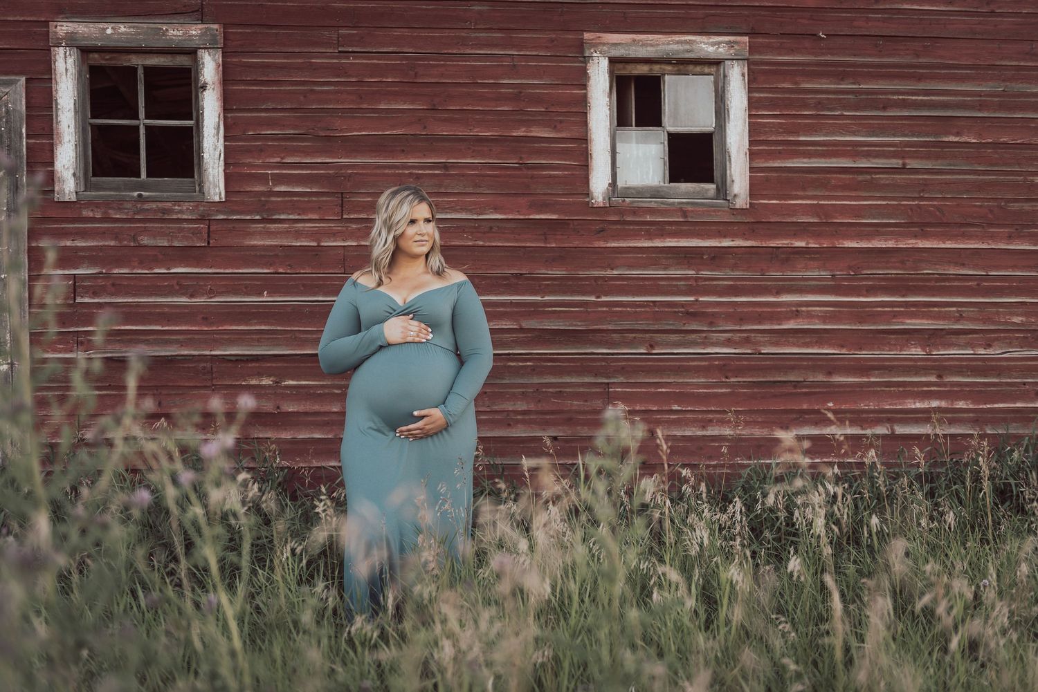 A maternity portrait against a rustic red barn surrounded by tall grass.