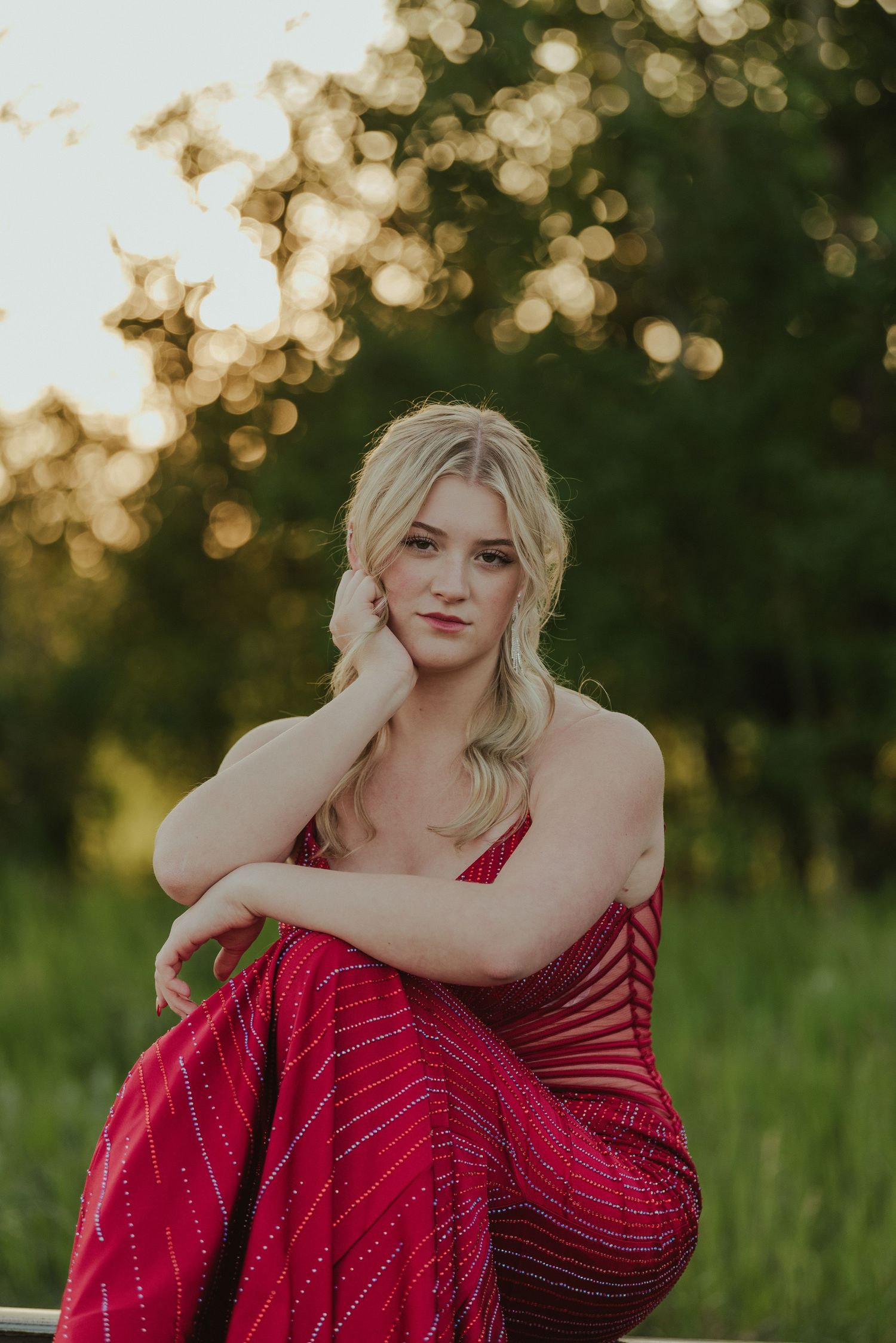 A person in a sparkly red dress poses outdoors during sunset with bokeh lighting.
