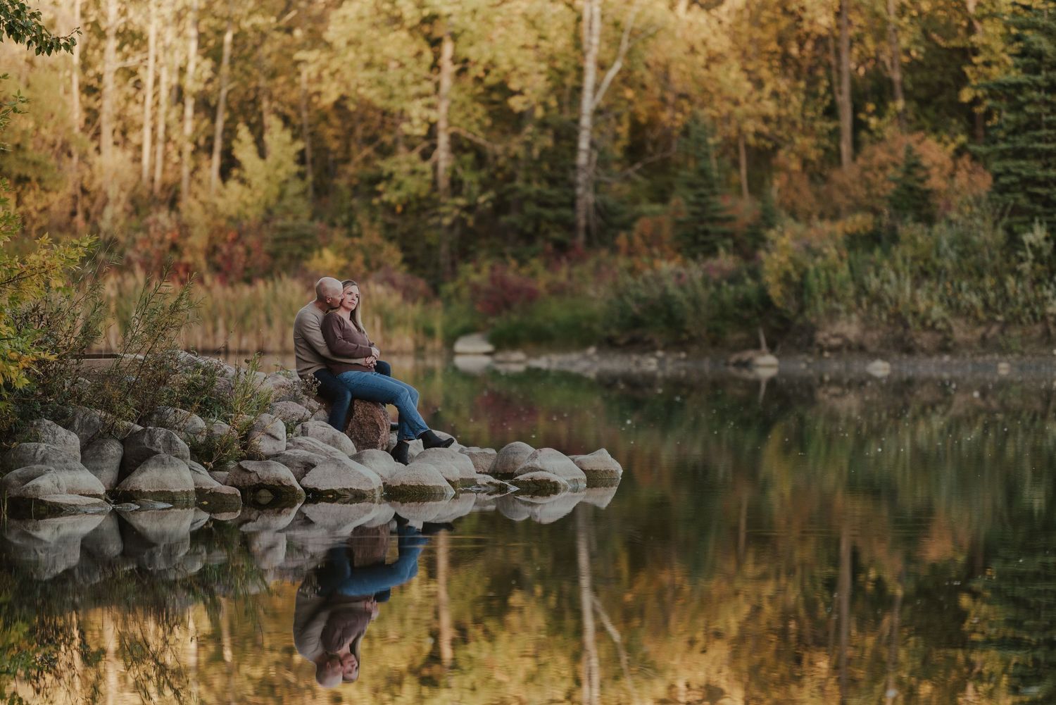 A tranquil autumn scene of someone fishing from a rocky shoreline reflected in calm lake waters at sunset.
