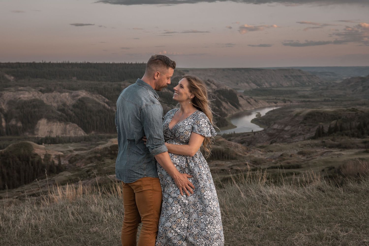 Couple embraces at sunset overlooking scenic canyon landscape with river winding through valley below.