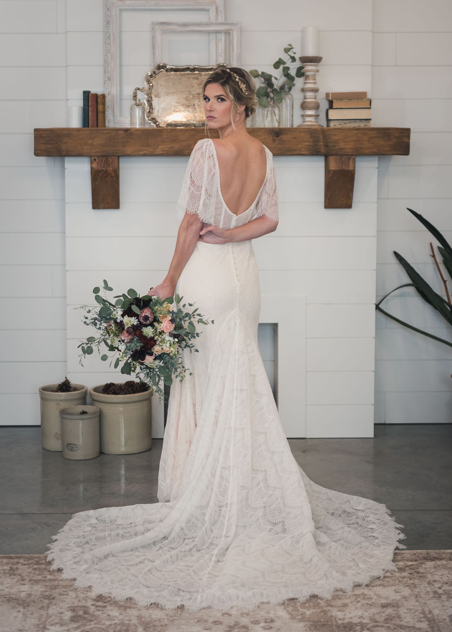 Elegant backless lace wedding dress with flowing train shown on model against white wall with rustic wooden shelves.