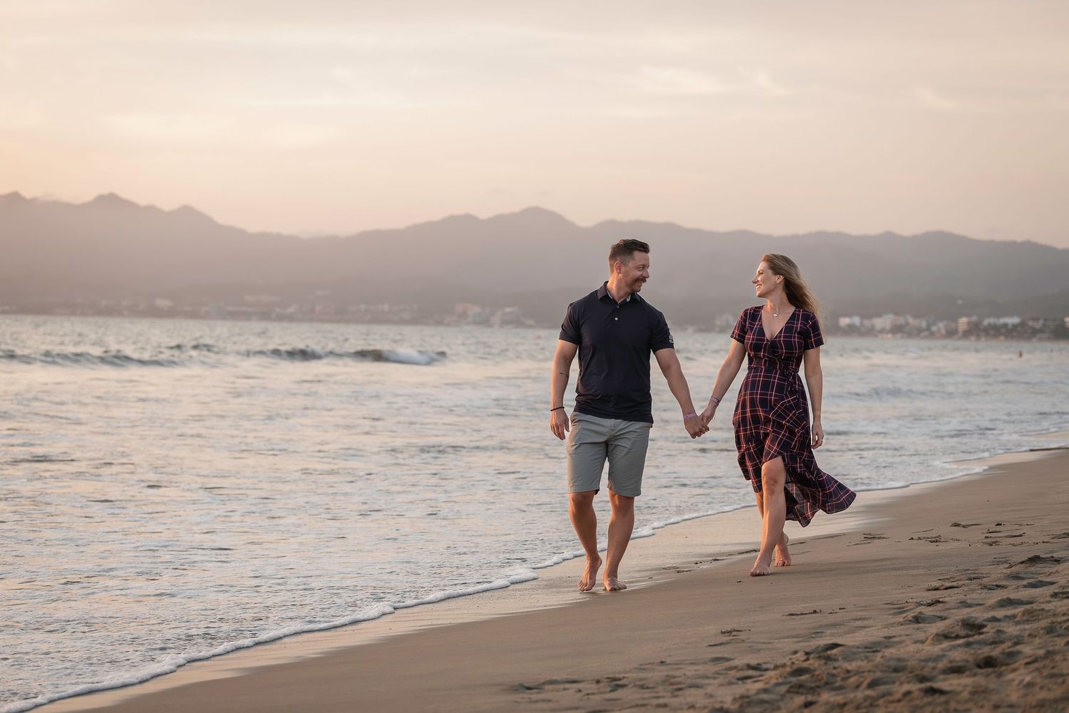 A couple walks hand in hand along a sandy beach with mountains and sunset in the background.
