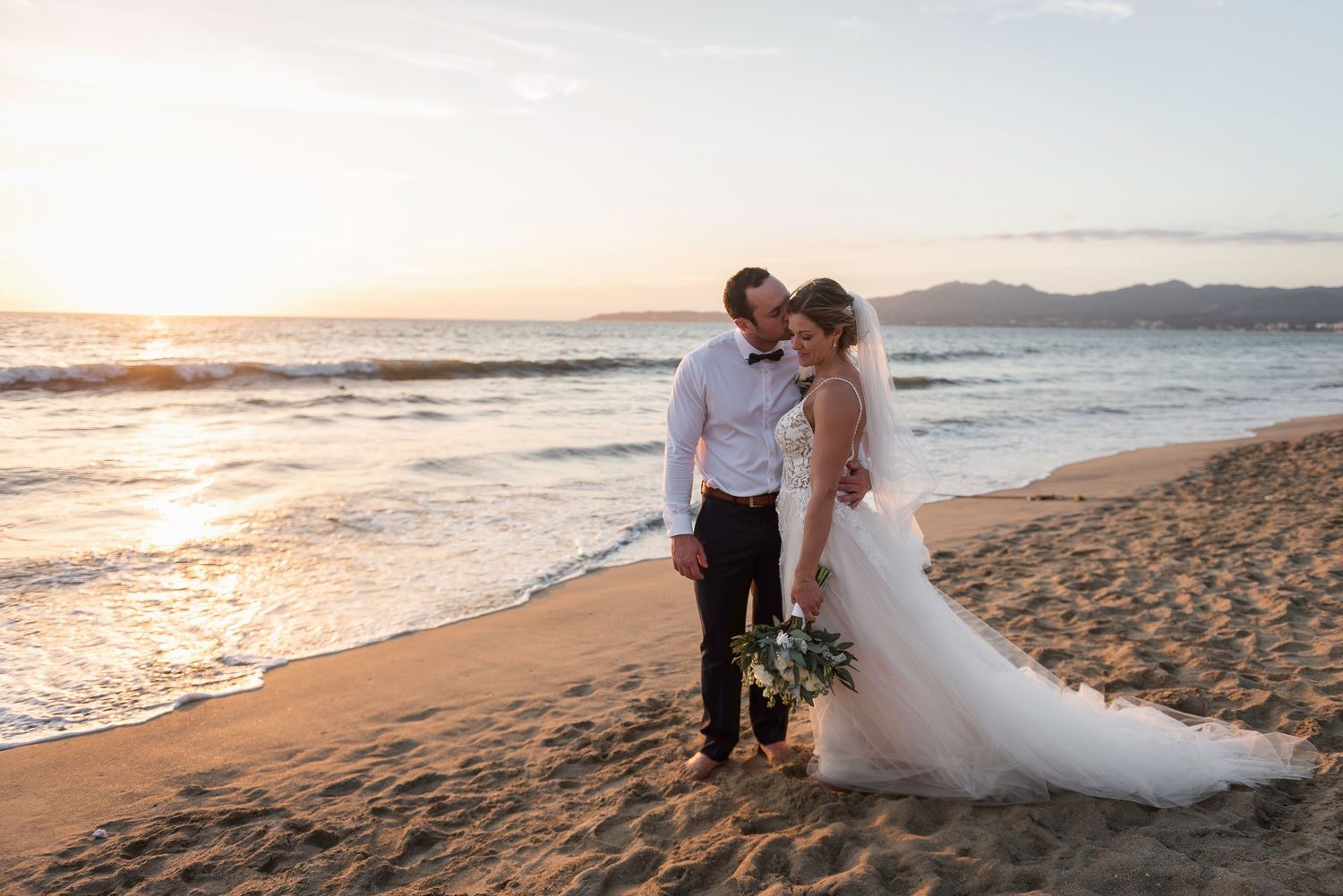 Newlyweds embracing on beach at sunset with waves and mountains in background.
