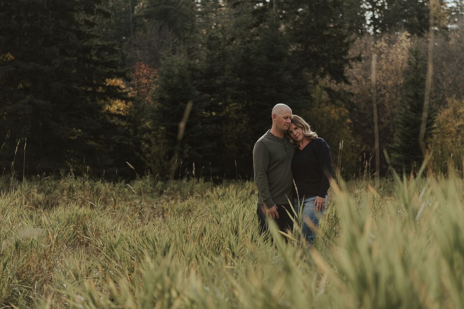 Couple sharing tender moment while walking through tall grass with forest backdrop.