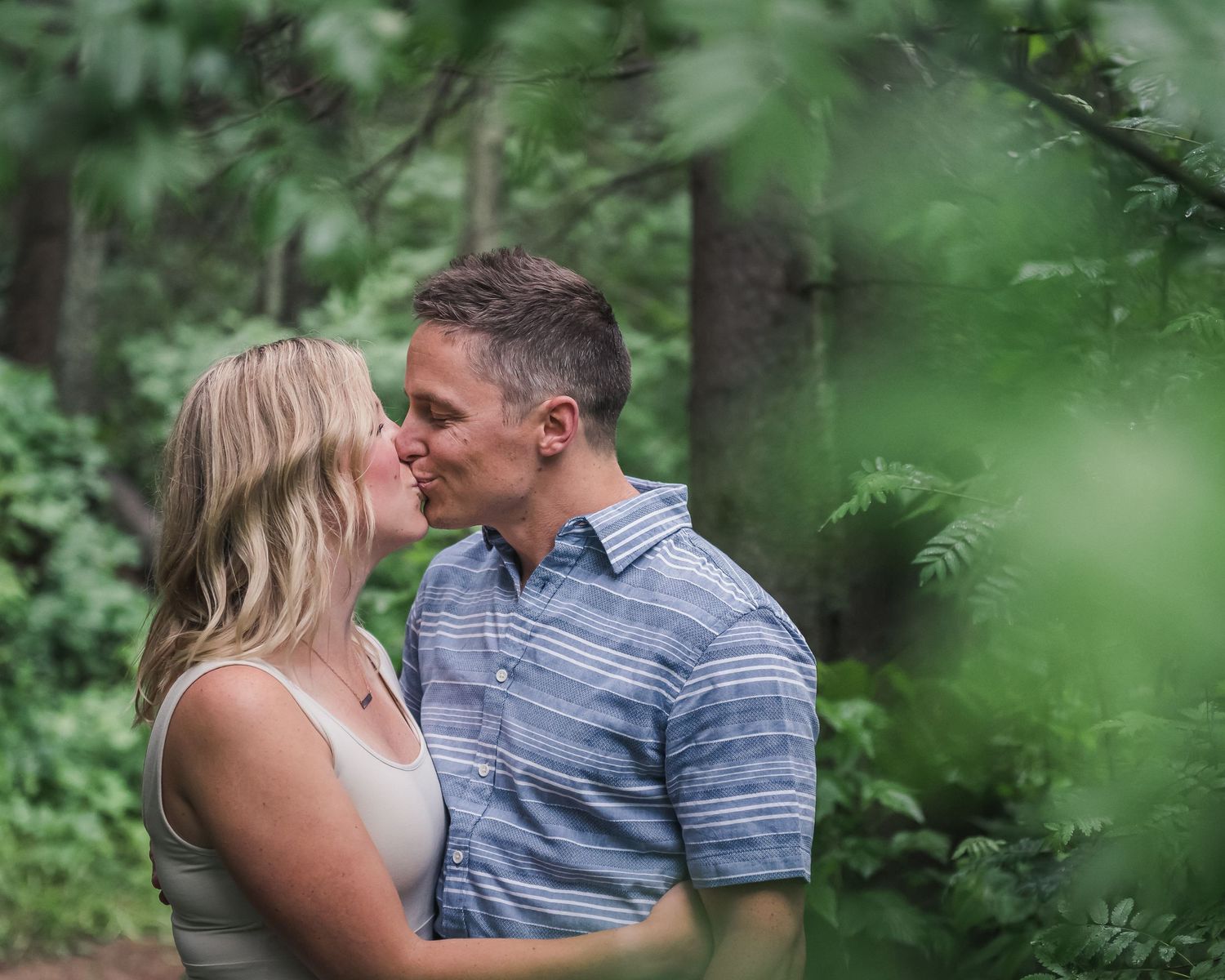 A romantic couple shares an intimate moment surrounded by lush green foliage in a forest setting.