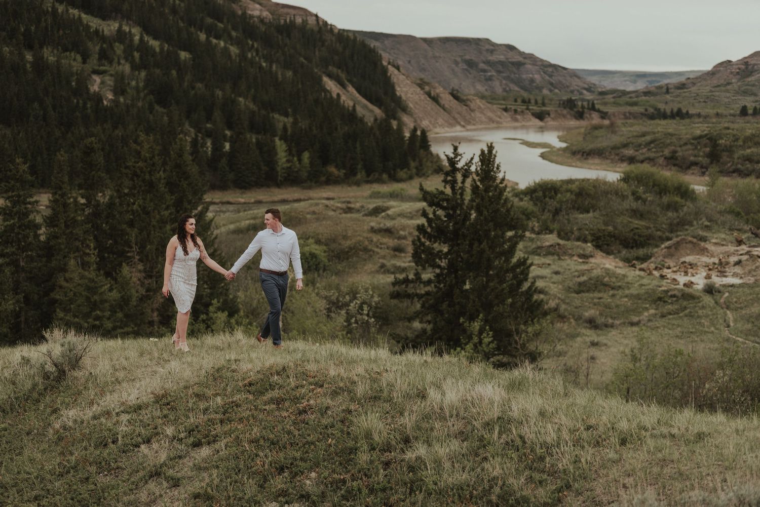 Couple holding hands while walking through mountainous terrain with river vista in background.