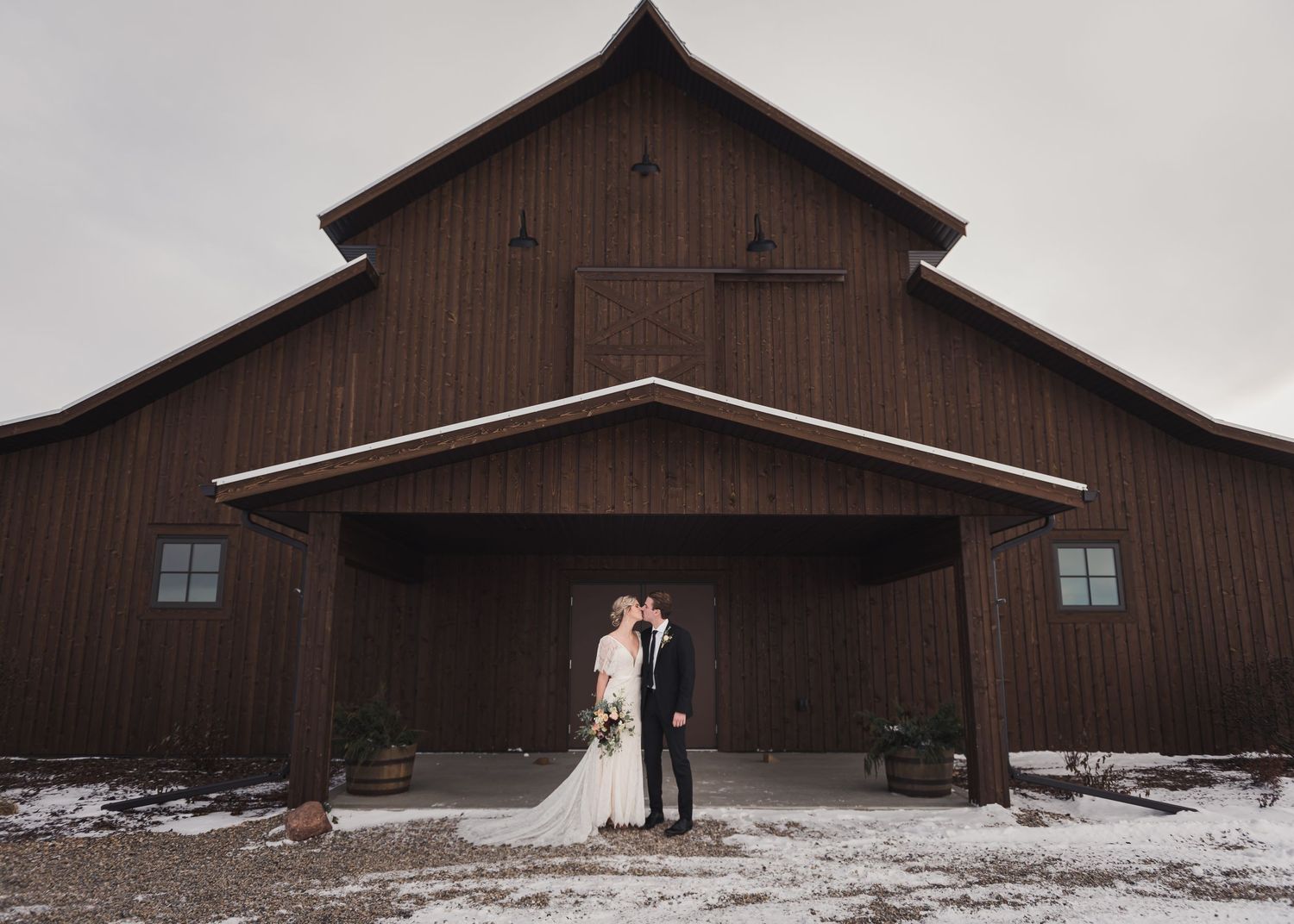 Newlyweds pose in front of rustic brown barn in winter setting.