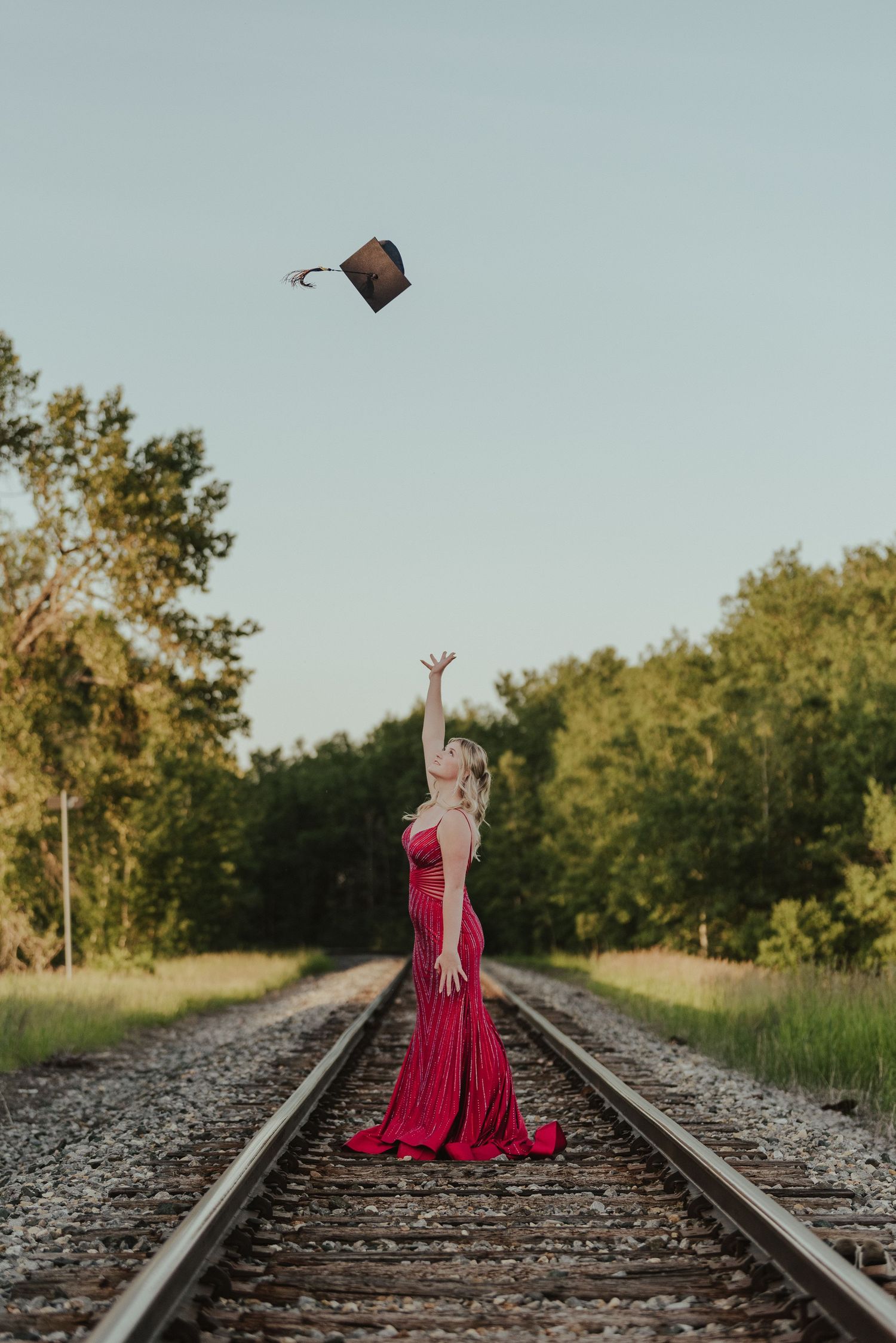 A figure in a red dress throws a graduation cap while standing on railroad tracks.