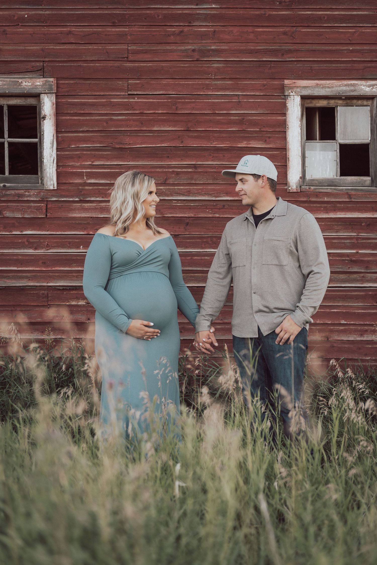 An expectant couple poses together by a weathered red barn in tall grass.