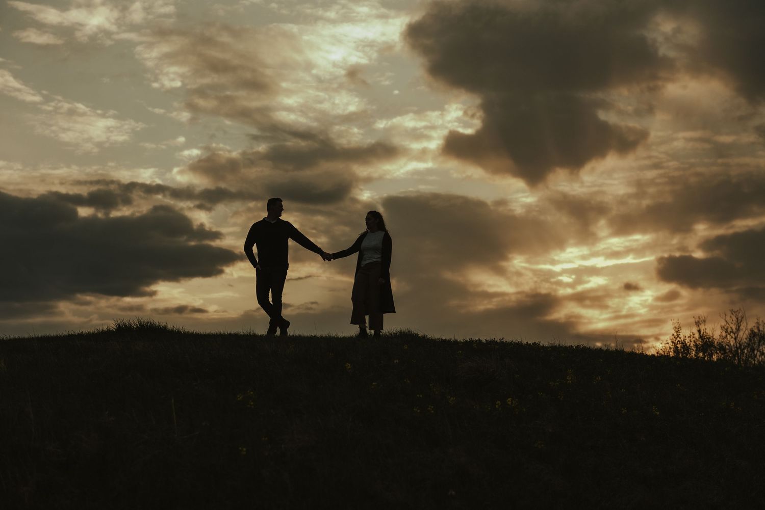Silhouetted couple running together on a hilltop against a dramatic sunset sky.