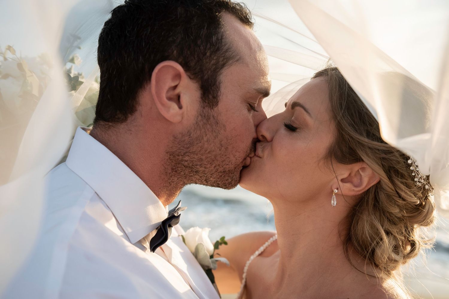 Newlyweds share a romantic kiss under a flowing white veil at their wedding ceremony.
