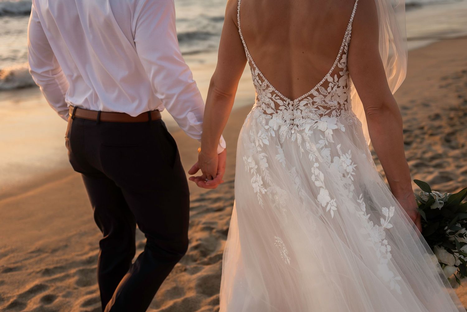 Close-up of bride's detailed lace backless dress as couple holds hands on sandy beach at sunset.