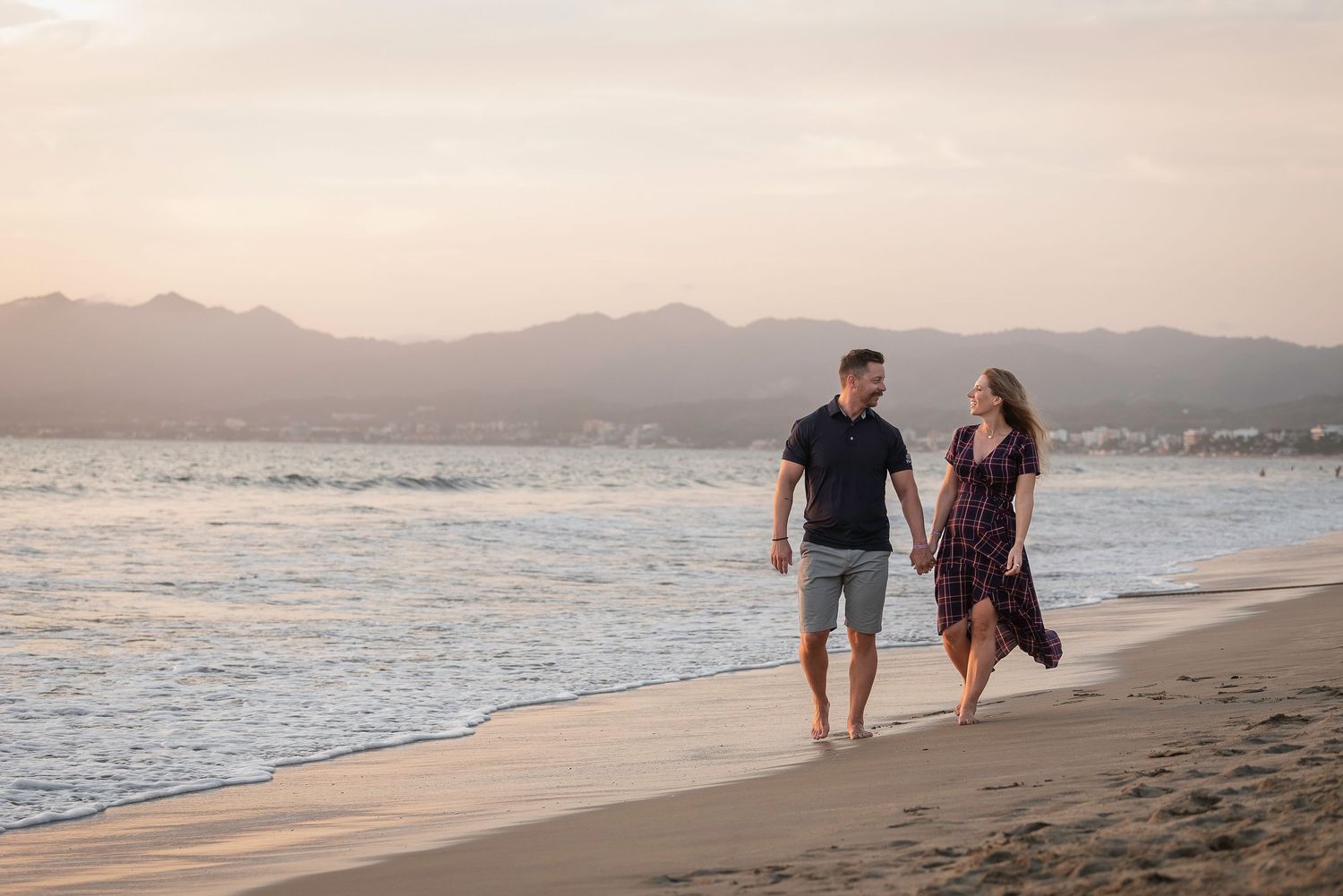 A couple walks hand in hand on a sandy beach at sunset with mountains visible in the background.