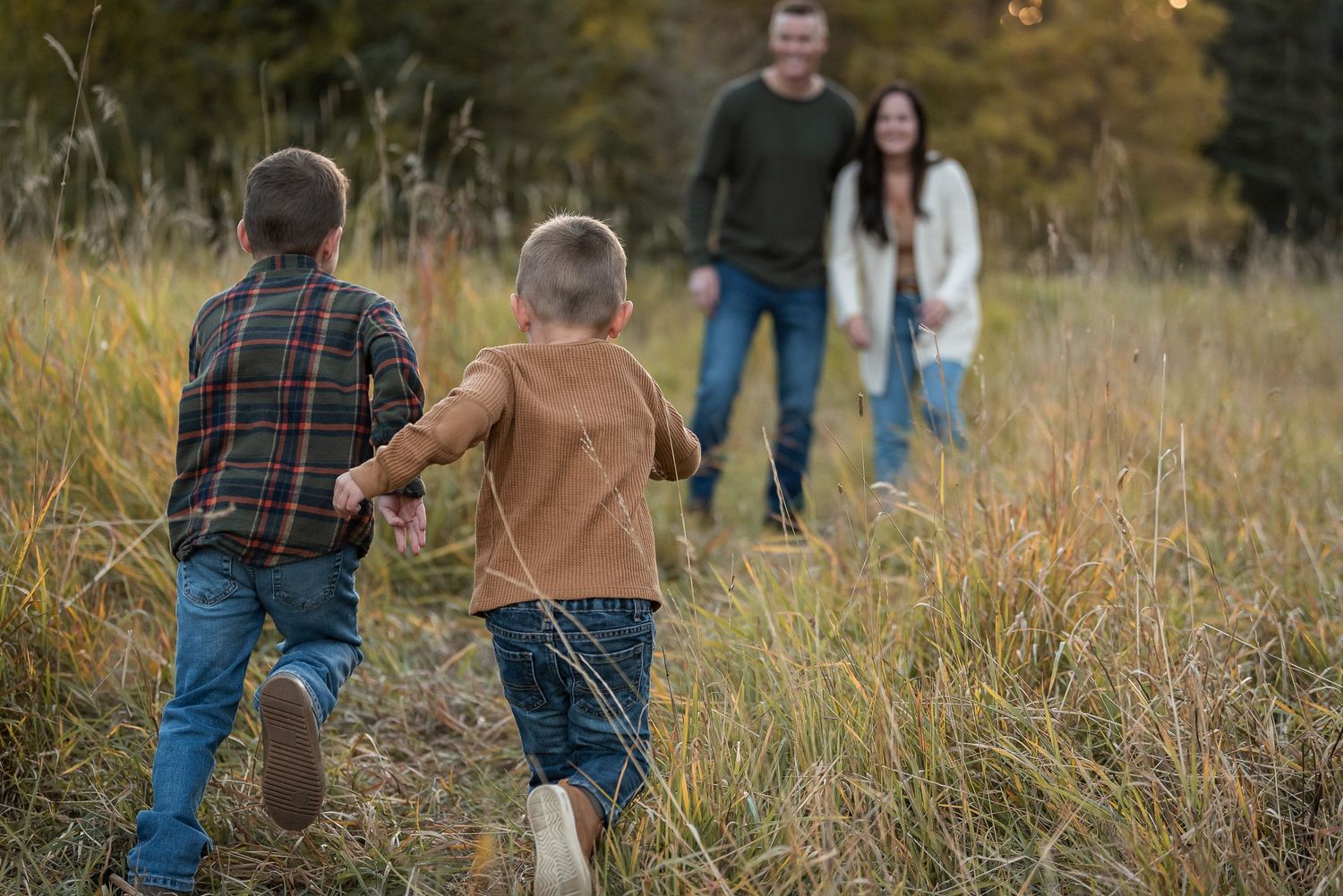 Two children run through tall autumn grass in a field during sunset with two adults watching from behind.