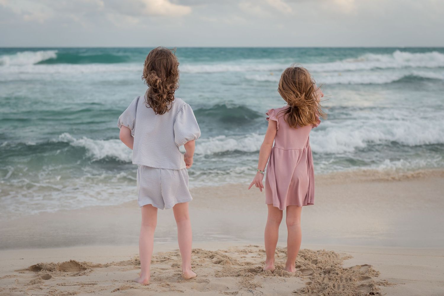 Two children in light colored outfits stand together on a beach at sunset watching the waves.