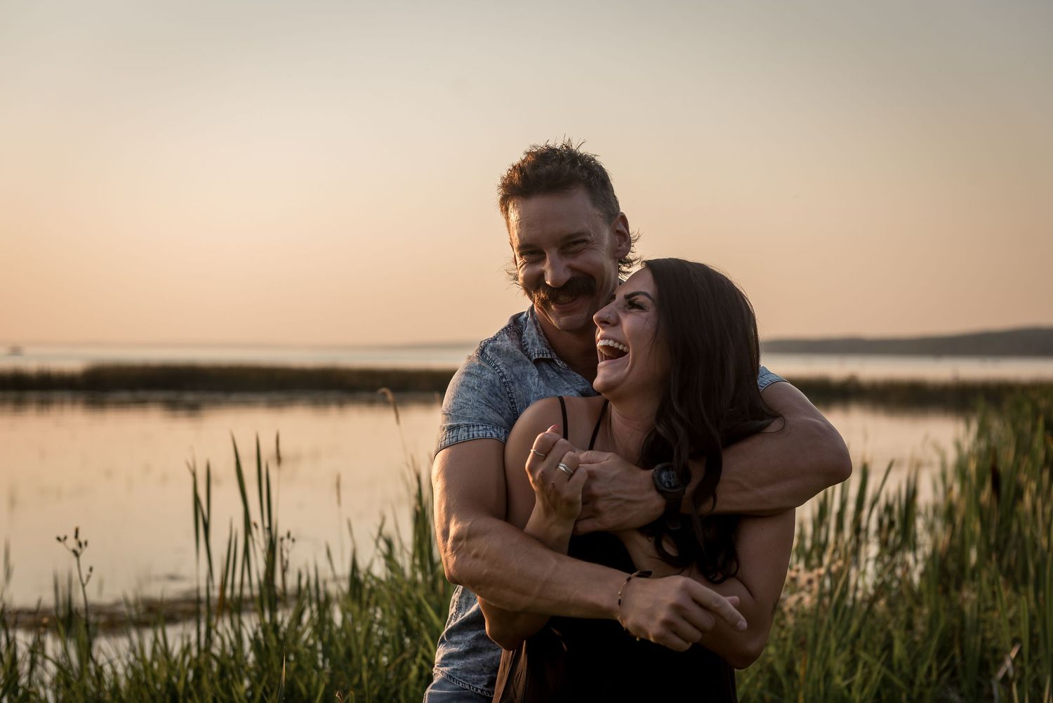 A romantic couple shares an intimate embrace at sunset near a tranquil lake with tall grasses in the foreground.