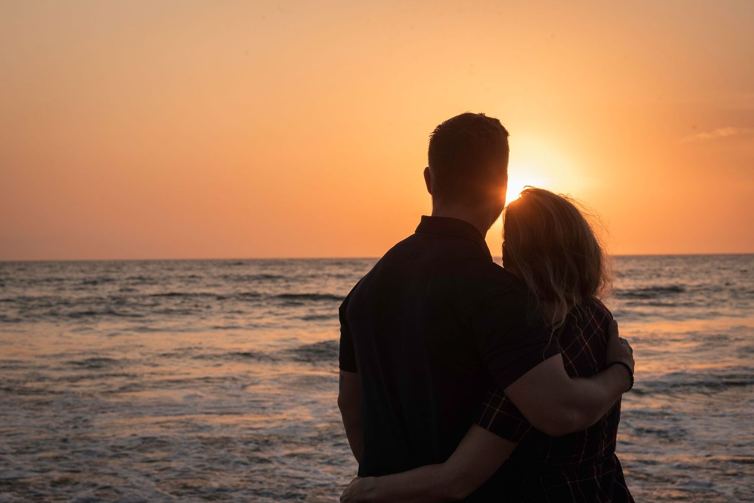 Silhouetted couple embracing while watching sunset over ocean waves.