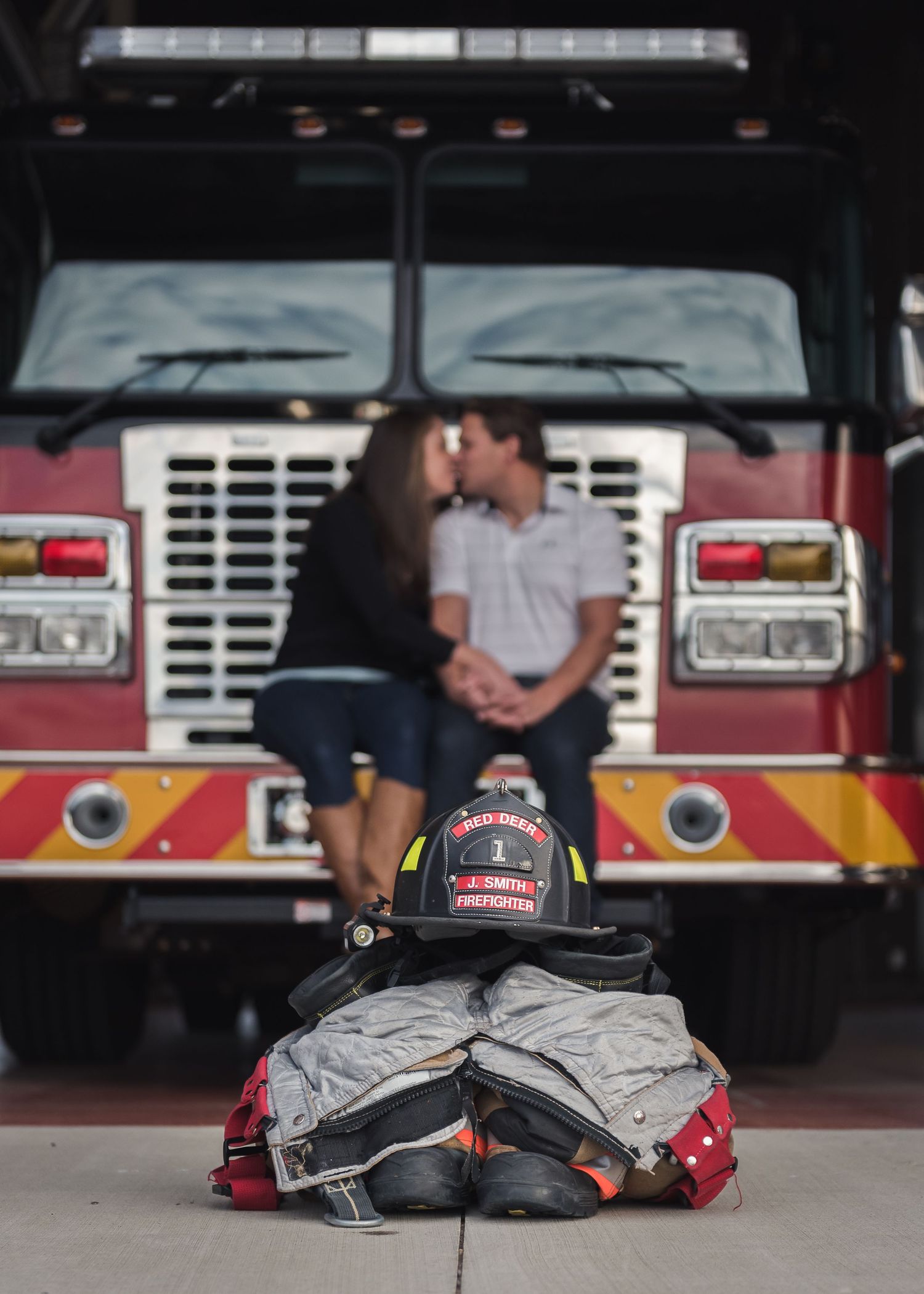 Firefighter gear and equipment arranged in front of a fire truck with couple in soft focus background.