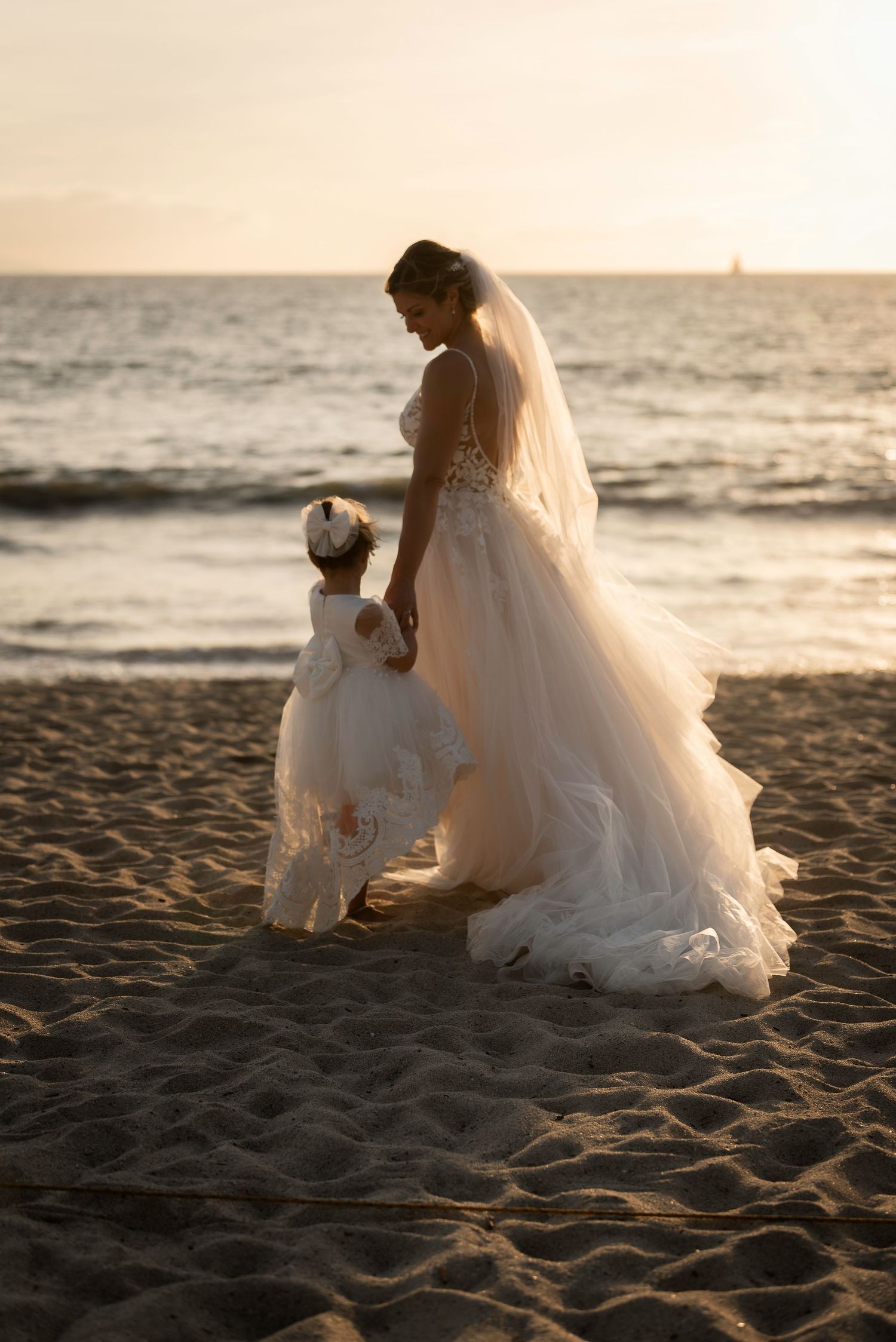 Bride in flowing wedding gown walks with flower girl on beach at sunset.