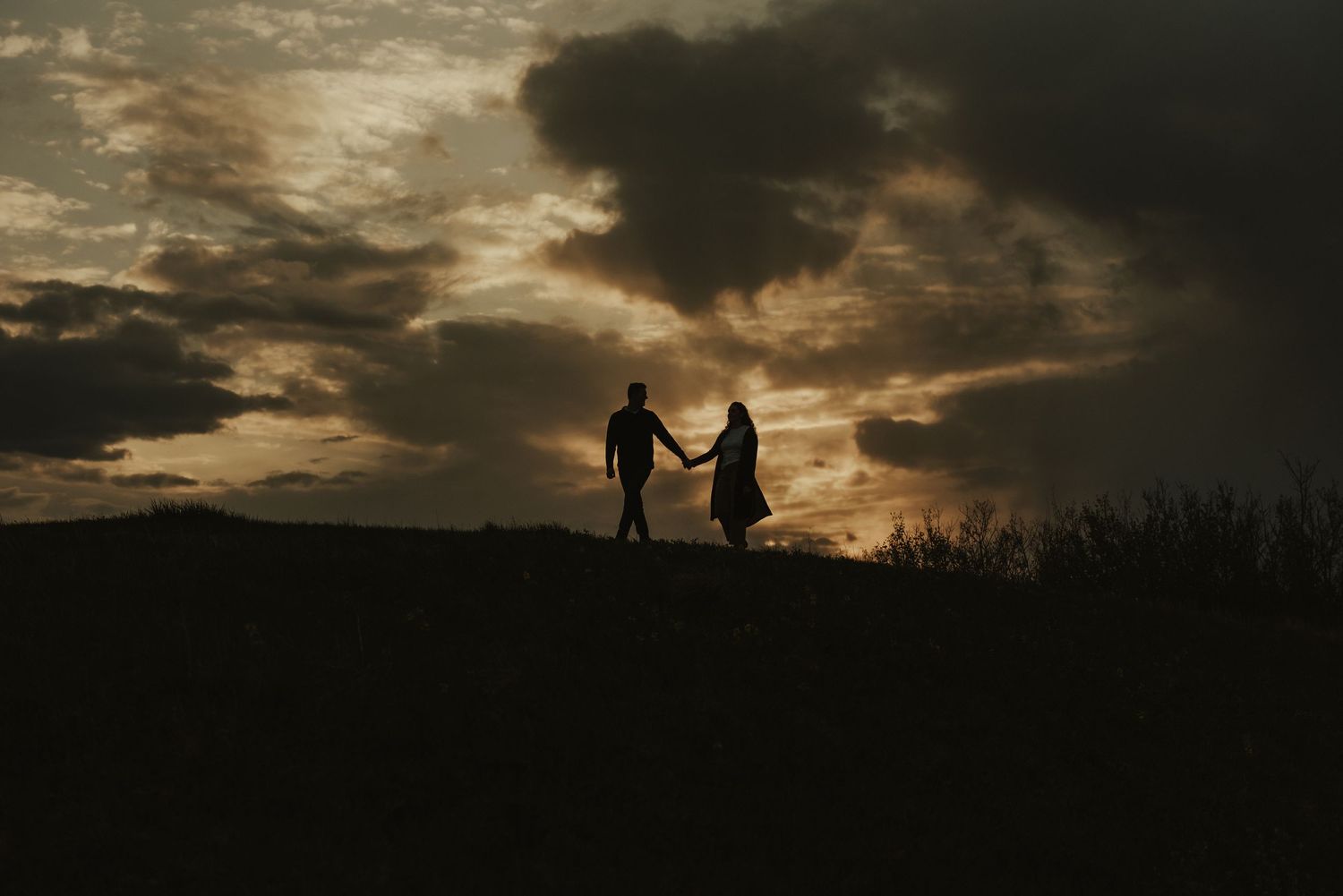Two silhouetted figures holding hands stand on a hilltop against a dramatic sunset with dark cloudy skies.
